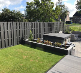 Een mooie antracieten schutting gecombineerd met een verhoogde plantenbak, steengrijze composieten vlonderplanken met een vierkante jacuzzi maken samen een mooie strakke tuin. Luxe antracieten schutting • antraciet gecoate dwarsbalken