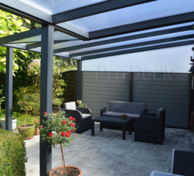 Grijze stapelplanken in combinatie met antracieten tuinpalen, gebruikt als wand bij een glazen overkapping. Perfect kleurend bij de overkappingskleuren. Stapelplankenschutting • grijs