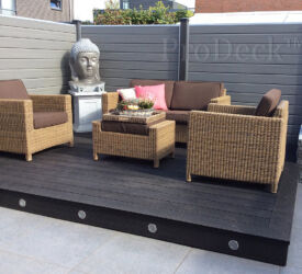 Een gezellige zithoek op een terras (met sfeerverlichting) van donkerantracieten vlonderplanken en een schutting van grijze stapelplanken met houtnerf. Composiet • vlonderplank • donkerantraciet • smalribbel • verhoogd vlonder